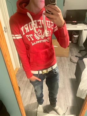 Röd Superdry hoodie med tryck - Snygg röd hoodie från Superdry med vitt tryck framtill, texten 'Superdry Sportswear 1977' och vita ränder på ärmarna. Hoodien har huva med vita snören och en stor magficka. Perfekt för en avslappnad och sportig stil.