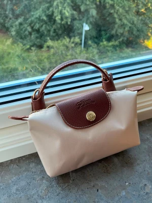 Beige och brun  handväska - Snygg beige handväska f med bruna detaljer i läder och guldfärgade metalldetaljer. Väskan har ett klassiskt vikbart design, korta handtag. Perfekt för dig som gillar stilrena accessoarer.