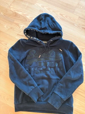 Svart hoodie från Superdry - Snygg svart hoodie från Superdry Icons med broderad vintage logo och japansk text på bröstet. Hoodien har dragsko med detaljer i guld och en stor magficka. Tillverkad i mjuk bomullsblandning och har ribbade muddar vid ärmar och nederkant.