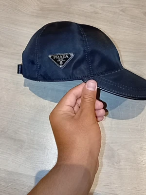 Prada svart keps - Snygg svart keps från Prada i nylonmaterial. Klassisk modell med böjd skärm och justerbar rem bak. Pradas ikoniska metallogo på sidan. Passar både dam och herr. Perfekt accessoar för en stilren och exklusiv look.