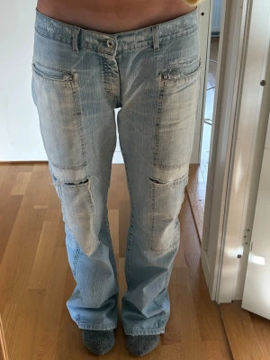Ljusblå cargojeans med slitningar - Säljer ett par ljusblå cargojeans med flera fickor och slitna detaljer både fram och bak. Jeansen har raka ben och en avslappnad passform. Materialet är klassisk denim i bomull och de har coola sömmar och patchdetaljer. Innerbenslängd: 83cm Midjemått rakt över: 41cm