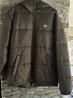 Levis jacka - Detta är en svart vadderad jacka med huva. Den har horisontella sömmar och två framfickor. Jackan stängs med dragkedja och har en liten Levi's-logotyp på bröstet med blå, vita och röda ränder. 