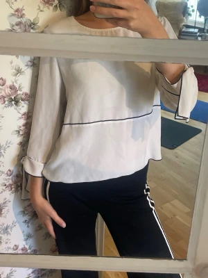 Vit blus med svarta detaljer - Säljer en stilren vit blus med svarta tunna linjer och knytband vid ärmsluten. Blusen har rund halsringning och är tillverkad i ett lätt, flödande material som ger en avslappnad men chic look. Perfekt att matcha med både jeans och kjol.