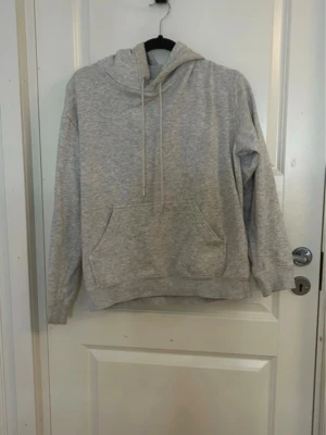 Ljusgrå hoodie med huva och ficka - En enkel och stilren ljusgrå hoodie med huva och dragsnören. Hoodien har en stor känguruficka framtill och ribbade muddar vid ärmslut och nederkant. Perfekt för en avslappnad och bekväm stil.