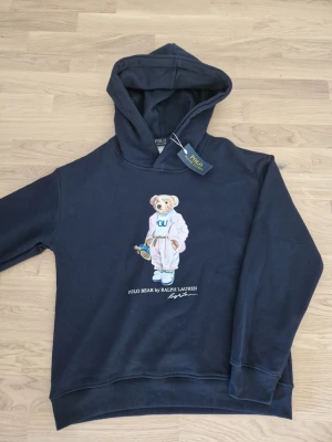  Polo Bear Hoodie  - Snygg marinblå hoodie från Polo Ralph Lauren med den ikoniska Polo Bear-figuren på bröstet. Hoodien har huva och ribbade muddar vid ärmar och nederkant. Tillverkad i mjuk bomull för skön komfort. Perfekt för en avslappnad och trendig stil. Ny med prislapp