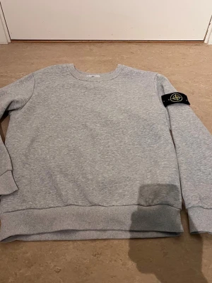 Grå sweatshirt från Stone Island - Snygg grå sweatshirt från Stone Island med klassisk rund hals och ribbade muddar. Tröjan har den ikoniska svarta patchen med gult och grönt Stone Island-märke på vänster ärm. Perfekt för en clean och stilren look.