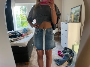 Blå jeanskjol med råa kanter - Säljer en ascool blå jeanskjol med råa, fransiga kanter och slitningar. Kjolen har en vit dekorativ rand framtill och klassiska fickor både fram och bak. 