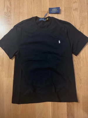Svart t-shirt från Polo Ralph Lauren - Klassisk svart t-shirt från Polo Ralph Lauren med rund halsringning och den ikoniska logon broderad i vitt på bröstet. Tillverkad i mjuk bomull för en skön känsla och perfekt passform. Enkel och stilren design som funkar till allt.