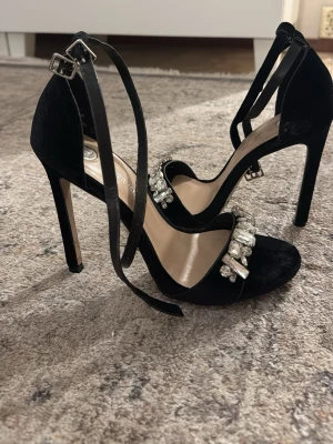Svarta pumps med strassdetaljer - Säljer ett par svarta pumps från River Island med höga smala klackar och rem runt ankeln. Skorna har öppen tå och är dekorerade med stora, glittrande strassdetaljer över foten. Perfekta för att lyfta vilken outfit som helst.