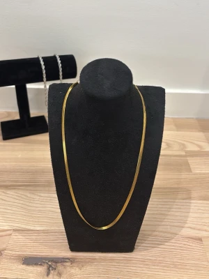 Guld halsband  - Snyggt och stilrent halsband i tunn, platt guldfärgad metall. Halsbandet har en enkel och elegant design med justerbar kedja och karbinlås. Perfekt för dig som gillar minimalistiska accessoarer och vill addera lite extra shine till din outfit.