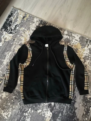 Svart hoodie med rutiga detaljer - Svart hoodie med dragkedja och klassiska rutiga paneler längs ärmar och axlar. Utrustad med huva och snören samt två stora fickor framtill. Perfekt för en chill och stilren look. Materialet känns mjukt och bekvämt.