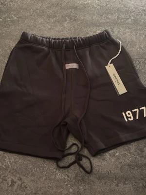 Svarta shorts Essentials 1977 - Snygga svarta shorts från Essentials med trycket '1977' på vänster ben. Shortsen har snörning i midjan och en relaxed passform. Perfekta för en chill och stilren look. Materialet känns mjukt och bekvämt, troligen bomull eller bomullsblandning.