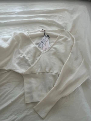 Vit stickad crop top med v-ringning - Trendig vit stickad crop top med bred ribbad midja och djup v-ringning. Långa ärmar och mjukt material som känns skönt mot huden. Perfekt att matcha med höga jeans eller kjol för en clean och stilren look.