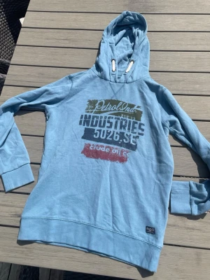 Ljusblå hoodie med tryck - Säljer en ljusblå hoodie med huva och vitt snöre. Framsidan har ett stort tryck i grönt, blått och rött med texten 'Petrol Ind. Industries 5026.SE crude oil'. Muddar vid ärmslut och nederkant. Perfekt för en avslappnad streetwear-look. Hör av dig vid frågor! 