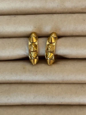 Guldiga hoops med nitar - Säljer ett par coola örhängen i form av små hoops i guldfärgad metall. Örhängena har flera spetsiga nitar runt hela ringen som ger en edgy och trendig look. Perfekta för dig som vill sticka ut och addera lite attityd till din stil.
