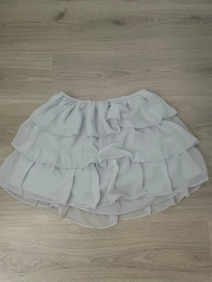 Vit volangkjol med resårmidja - Söt vit kortkjol med flera lager volanger och elastisk midja. Kjolen har en luftig och flowy känsla, perfekt för varma dagar. Tillverkad i ett lätt material som ger en romantisk look.