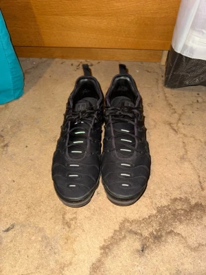 Svarta Nike VaporMax Plus sneakers - Säljer ett par svarta Nike VaporMax Plus sneakers med klassisk vågig design och reflekterande detaljer på ovandelen. Skorna har en robust sula med synliga luftkuddar och snörning framtill. Perfekta för dig som gillar streetwear och sportig stil.