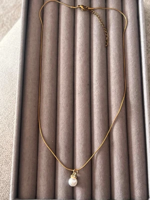 Guldhalsband med pärlhänge - Snyggt halsband i guldton med en rund vit pärla som hänge. Kedjan är tunn och elegant, och pärlan är dekorerad med en liten rad gnistrande stenar ovanför. Perfekt för dig som gillar stilrena accessoarer med en touch av lyx.