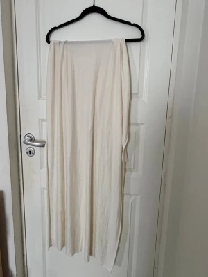 Krämvit lång sjal i mjukt tyg (jersey) - Säljer en krämvit, lång och rektangulär sjal i tunt och mjukt material. Perfekt att svepa runt halsen för en clean och stilren look. Enkel design utan mönster och passar till många olika stilar.