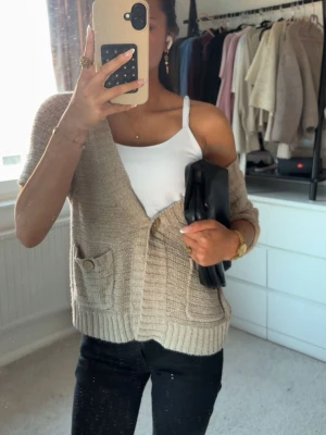 Beige kortärmad stickad kofta - Kortärmad beige stickad kofta med två fickor framtill, båda med knappdetaljer. Koftan har v-ringning, ribbade muddar och stängs med två träknappar. Perfekt att slänga över ett linne för en avslappnad look💞