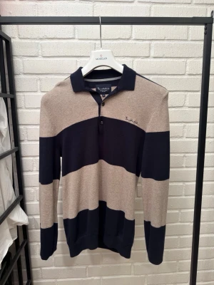 Randig pikétröja från Bondelid - Snygg långärmad pikétröja från Bondelid i beige och marinblått med breda horisontella ränder. Tröjan har krage, knäppning vid halsen och diskret logga broderad på bröstet. Perfekt för en stilren och avslappnad look.