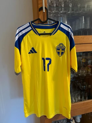 Sverige matchtröja nr 17 Gyökeres - Gul och blå Sverige matchtröja från Adidas med nummer 17 och namnet Gyökeres på ryggen. Tröjan har korta ärmar, blå detaljer på axlarna och det svenska landslagets emblem på bröstet. Perfekt för fotbollsfans!