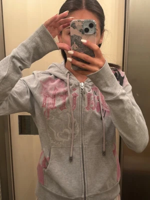 Grå hoodie med rosa detaljer och dragkedja - Säljer en grå hoodie med dragkedja och huva, dekorerad med rosa flames och tryck både fram och bak. Hoodien har fickor på sidorna och långa ärmar med tumhål. Materialet ser ut att vara mjukt och bekvämt, perfekt för en chill look.