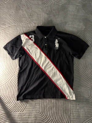 Svart ralphlauren piké - Svart pikétröja med vit diagonal rand och röda detaljer. Tröjan har en stor broderad logga på bröstet och även en trea på andra sidan. Tröjan är i storlek L och passar perfekt på ungefär 180-190 beroende på hur du vill att den ska sitta. Fräsch tröja utan synliga defekter. I kragen står det även Ralph lauren. Har även fler liknande ralphlauren pikéer i andra storlekar och färger på min profil så gå in och kika vetja! Och såklart om man köper fler blir priset billigare 😉Skriv vid funderingar 👍