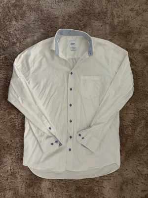 Vit skjorta från Eton of Sweden - Snygg vit skjorta från Eton of Sweden med blåmönstrad insida på kragen och blå knappar. Skjortan har en bröstficka och lång ärm. Fitted body-modell och tillverkad i bomull. Perfekt för dig som gillar stilrena detaljer.