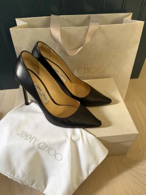 Svarta pumps från Jimmy Choo - Jimmy Choo Romy i svart skinn, stl 37. Klassisk modell med spetsig tå och stilettklack  (10 cm).   Använda med normalt slitage, främst på lädersulor, tåspetsar och mindre märken på klackarna (se bilder).   Originalkartong och dust bags medföljer.