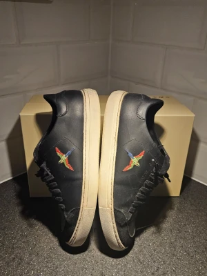 Svarta sneakers från Axel Arigato - Stilrena svarta sneakers från Axel Arigato med broderad färgglad fågel på sidan. Skorna har svarta skosnören och vit sula. Perfekta för dig som gillar minimalistisk design.