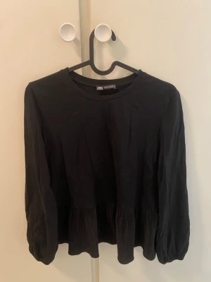 Svart blus med plisserad nederdel Zara - Svart blus från Zara med rund halsringning och långa ärmar. Blusen har en plisserad detalj nedtill som ger en snygg volang-effekt. Tillverkad i ett tunt, lätt material som känns skönt mot huden. Enkel men trendig design som passar till mycket.