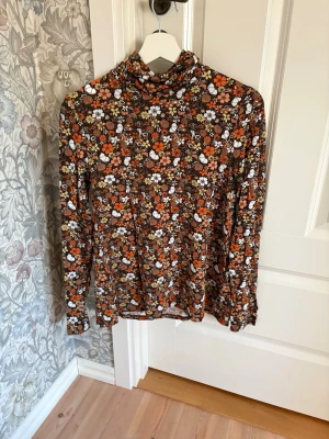 Blommig polotopp från Jumperfabriken - Säljer en tunn och stretchig polotopp från Jumperfabriken i storlek S. Toppen har ett färgglatt blommigt mönster i brunt, orange, vitt och gult samt lång ärm och hög krage. Perfekt för dig som gillar retrovibbar och vill sticka ut lite extra.
