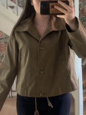 Trenchcoat  - Snygg jacka perfekt till våren eller hösten med en stickad under. Färgen är Khaki färg eller än grön/brun. Använd ett par gånger! 
