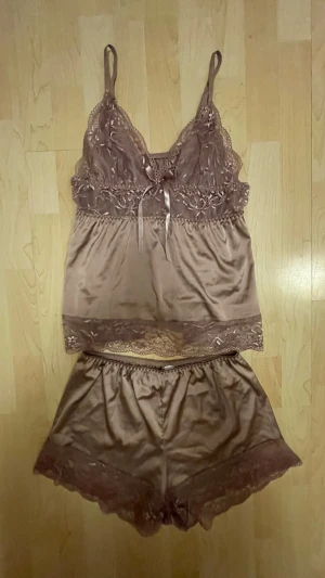 Rosa satinpyjamas med spetsdetaljer - Säljer ett set med linne och shorts i skimrande rosa satin. Linnet har tunna axelband, spets vid bysten och en liten rosett framtill. Shortsen har matchande spetskant nedtill. Båda delarna är mjuka och har en lyxig känsla.