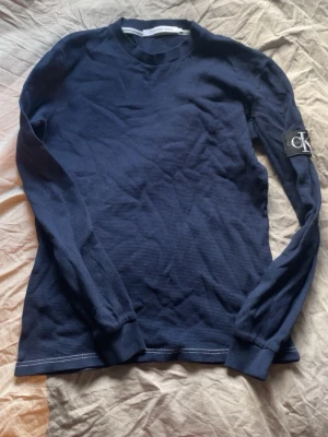 Mörkblå långärmad tröja CK Jeans - Snygg mörkblå långärmad tröja från Calvin Klein Jeans med rund halsringning och ribbade muddar. Tröjan har en tydlig CK-logga på ärmen och är tillverkad i ett strukturerat, mjukt material som känns skönt mot huden.