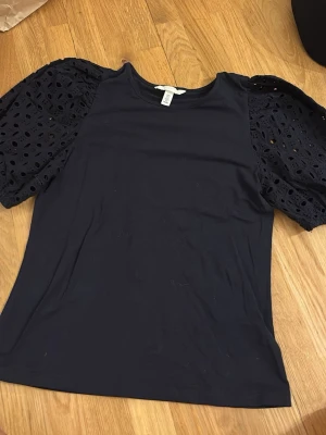 Svart topp med puffärmar från H&M - Säljer en svart topp från H&M med snygga puffärmar i broderad spets. Toppen har rund halsringning och är tillverkad i mjuk bomull. Ärmarna är korta och ger en unik look med sina håldetaljer. Perfekt för dig som vill sticka ut lite extra.