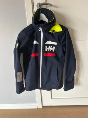 Marinblå vindjacka Helly Hansen - Snygg marinblå vindjacka från Helly Hansen med vit dragkedja, röda och vita detaljer samt reflex på ärmen. Jackan har huva med gul insida och tryckt logga på bröstet. Perfekt för blåsiga dagar och sportiga outfits.