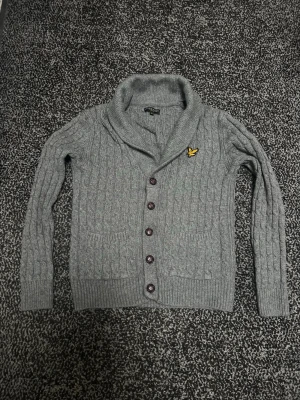 Grå vintage kofta lyle & scott  - Snygg grå kabelstickad kofta med sjalkrage och fem mörka knappar framtill. Koftan har ett gult broderat märke på bröstet och ribbade muddar vid ärmslut och nederkant. Perfekt för lager på lager och en stilren look. 