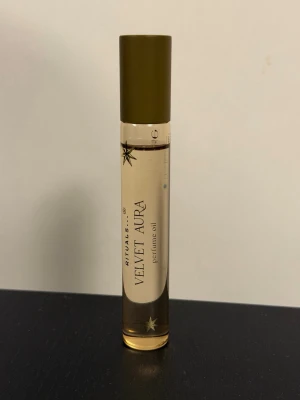 Rituals Velvet Aura perfume oil - Velvet Aura perfume oil från Rituals i en smal glasflaska med guldfärgat lock. Flaskan är transparent och visar den ljusgula oljan inuti. Elegant design med stjärndetaljer på etiketten. Perfekt att ha med i väskan för en fräsch doft när som helst.