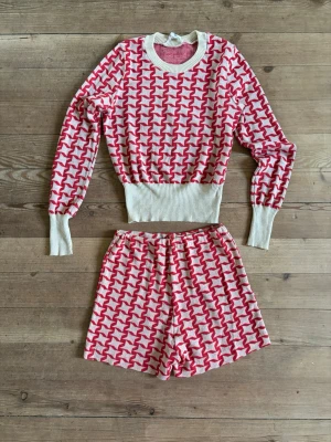 RETRO! Stickat set i rött och vitt mönster - Säljer ett stickat loungewear-set med tröja och shorts i vitt och rosa grafiskt mönster. Tröjan har bred ribbad mudd i midjan och vid ärmsluten samt rund hals. Shortsen har elastisk midja för extra komfort. Perfekt för chill dagar hemma. Set i stickat material. Jag skulle säga att det är S/M i strl. Shortsen slutar under rumpan. Resår i midjan. Snyggt på tennisplanen kanske 🎾.  Fläckarna på mudden vid halsen syns knappt. Har ej testat att tvätta bort så vem vet…