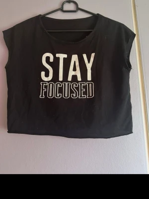 Svart croppad topp med tryck - Cool svart croppad topp med texten 'STAY FOCUSED' i vitt framtill. Toppen är ärmlös och har en avslappnad passform. Perfekt till jeans eller shorts för en casual look. Fint skick och knappt använd! Osäker på exakt storlek men skulle tippa på S eller M