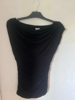 Svart topp från H&M med draperad hals - Säljer en svart topp från H&M i storlek S. Toppen har en snygg draperad halsringning och är figurnära med ett mjukt, stretchigt material. Perfekt att styla med jeans eller kjol för en trendig look 💗