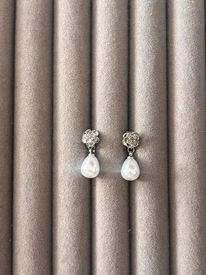 Silvriga örhängen med droppformade pärlor - Säljer ett par eleganta örhängen i silverfärg med små blommor upptill och droppformade vita pärlor som hänger ned. Perfekta för dig som gillar klassiska smycken med en romantisk touch. Stift och bakstycke i metall.