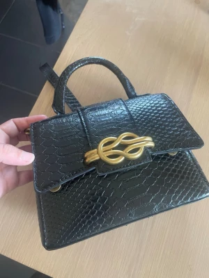 Svart handväska med ormmönster - Snygg svart handväska i ormmönstrat konstläder med guldfärgat dekorativt spänne framtill. Väskan har både handtag och axelrem med guldkedja, vilket ger en trendig look. Perfekt storlek för det viktigaste och passar till många outfits. Liknar B A L E N C I A G A väskan😃