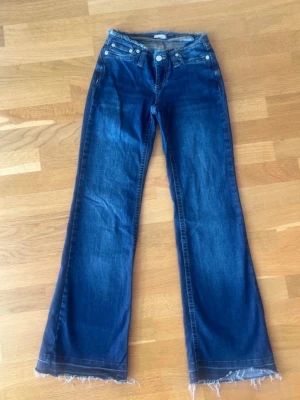 Blå bootcut jeans med rå kant - Säljer ett par mörkblå bootcut jeans med rå, fransig kant nertill. Jeansen har klassisk femficksmodell, låg midja och slitningar för en cool vintagekänsla. Perfekta för dig som gillar en avslappnad och trendig stil.