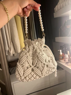Vit virkad handväska med pärlhandtag - Superfin handväska i vit virkad design med snyggt mönster och pärlhandtag. Väskan har dragskostängning och är perfekt för dig som gillar boho-stil och unika accessoarer. Materialet är mjukt och väskan känns både trendig och söt.