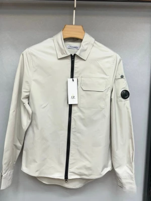 C.P. Company zip overshirt  - Stilren beige overshirt från C.P. Company med svart dragkedja, bröstficka och ikonisk linsdetalj på ärmen. Långärmad modell i ett lätt och slätt material, perfekt för lager-på-lager. Klassisk krage och modern passform.