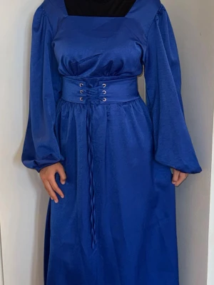 Blå långklänning med snörning - Snygg blå långklänning med markerad midja och bred snörning framtill. Klänningen har långa, puffiga ärmar och fyrkantig ringning. OBS! Modellen är XS och bär storlek L