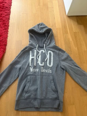 Grå hoodie med HCO-tryck - Snygg grå hoodie med stor huva och dragsko. På bröstet står det 'HCO Wave Devils' i stora vita bokstäver. Hoodien har en klassisk känguruficka framtill och är gjord i mjukt sweatshirtmaterial. Perfekt för en avslappnad och cool stil.